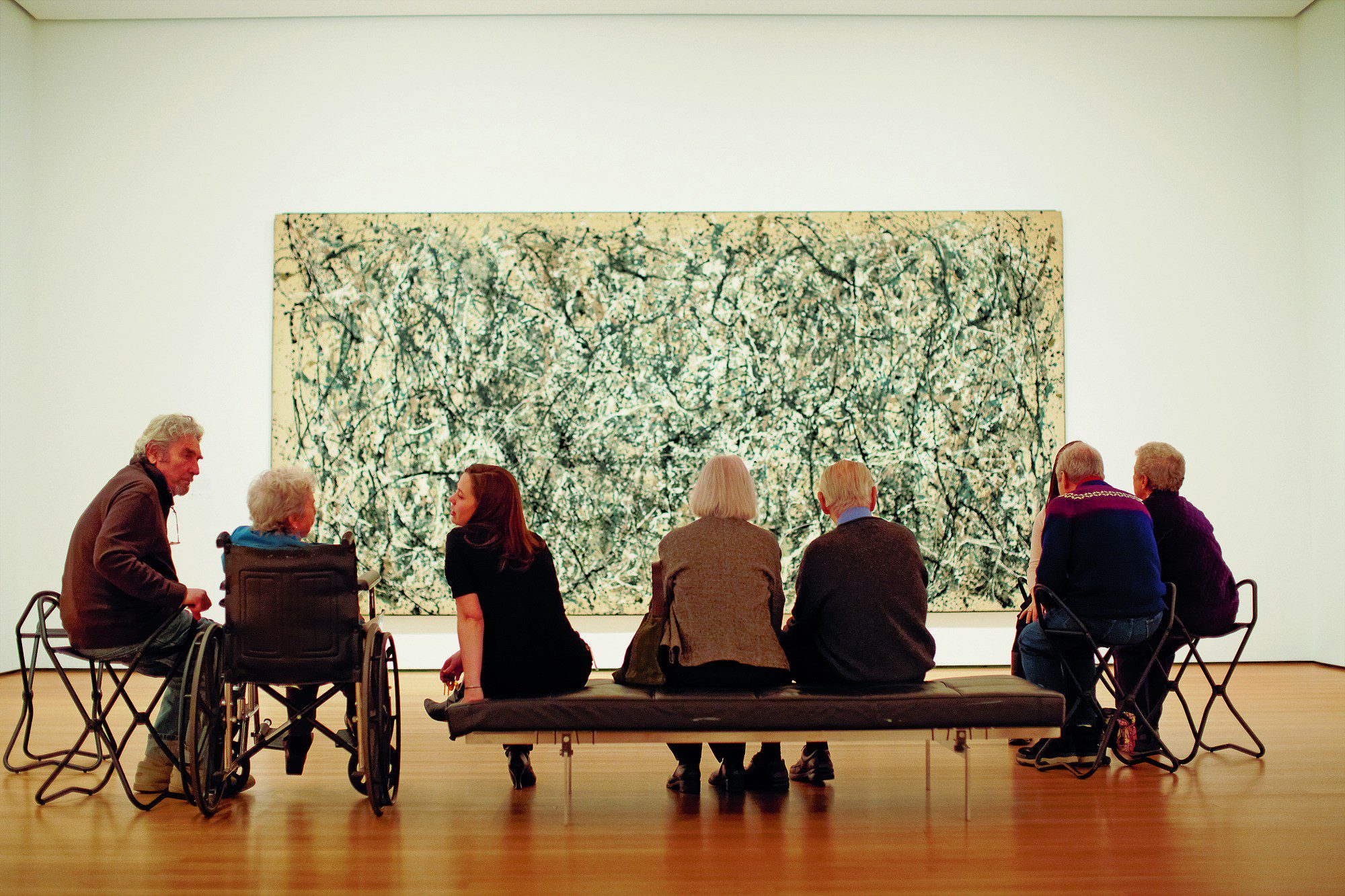 Seis personas de la tercera edad, algunos en silla de ruedas, se sientan frente a una obra de arte.